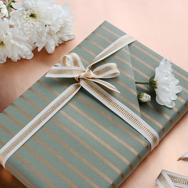 A gift wrapped with a striped sage/gol kraft wrapping paper with a folded pocket containing a white flower. Finished with a creme-gold ribbon.
